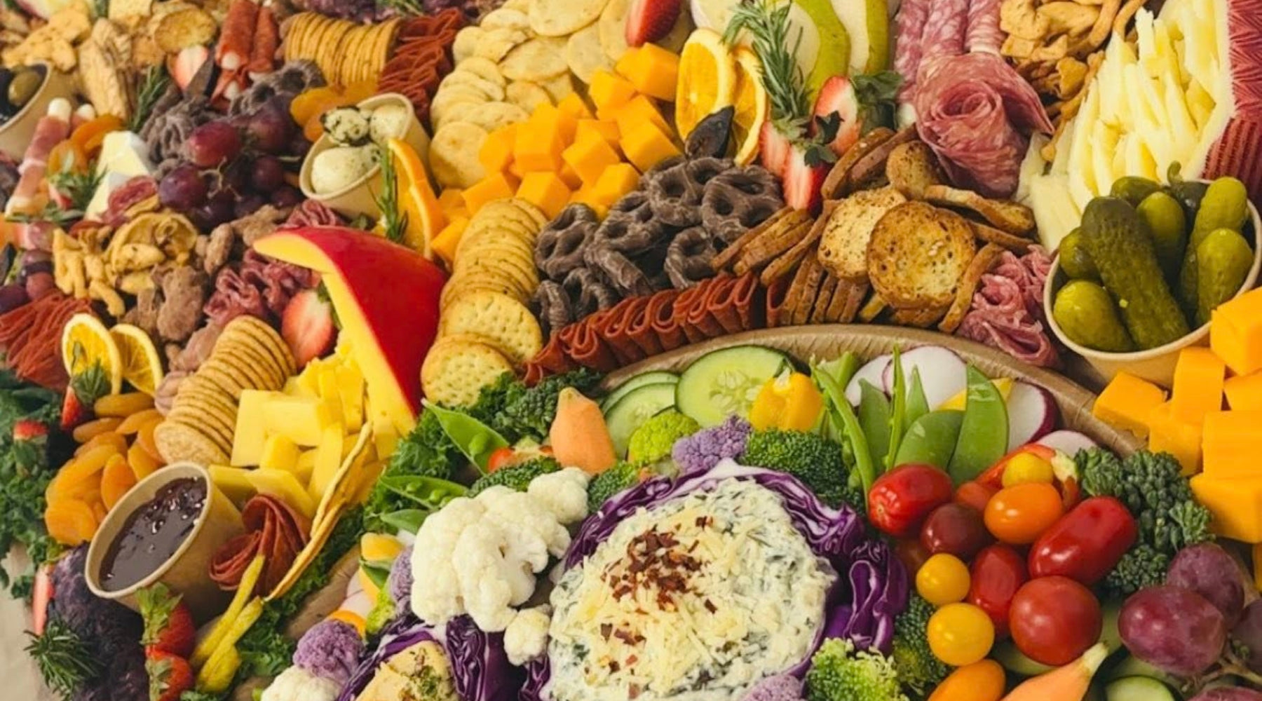 Close-up of a grazing table with cheeses, cured meats, crackers, pickles, fresh fruit, and veggie dip