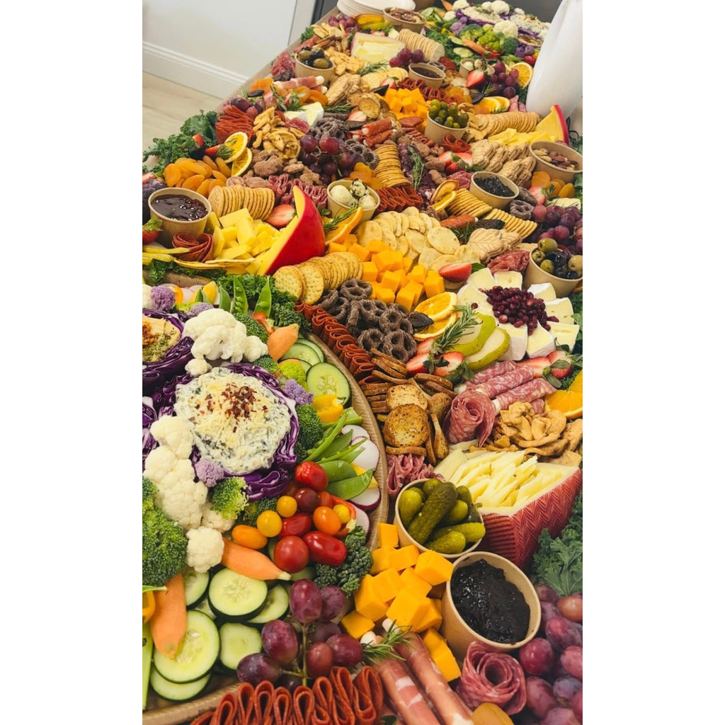 Close-up of a grazing table with cured meats, cheese cubes, crackers, grapes, olives, and a fresh vegetable platter