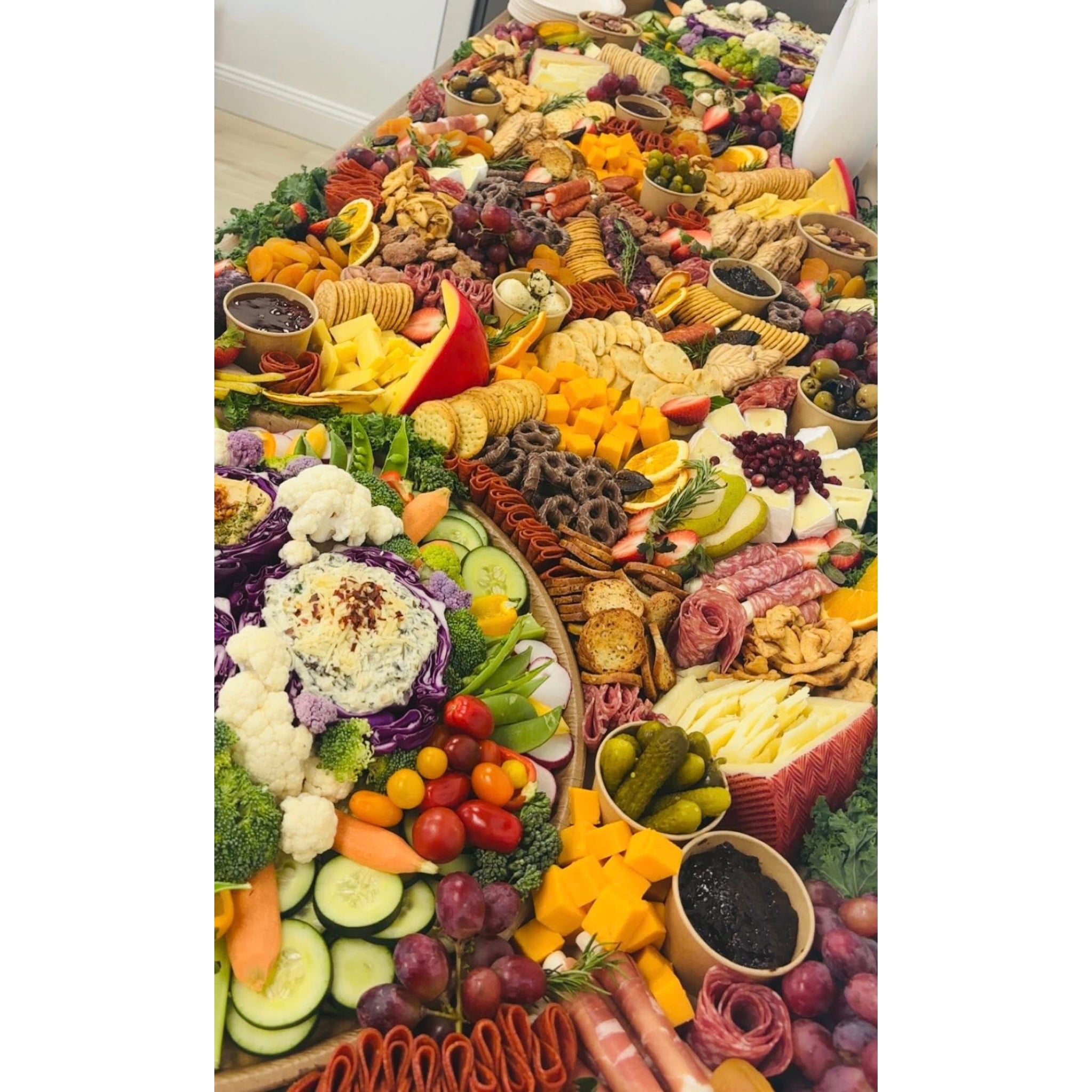 Close-up of a grazing table with cured meats, cheese cubes, crackers, grapes, olives, and a fresh vegetable platter